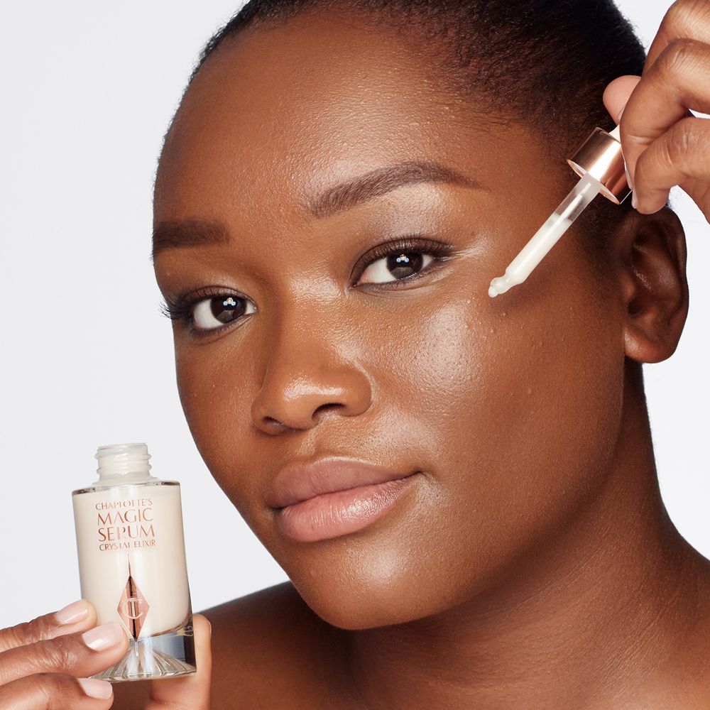 A deep-tone model with brown eyes wearing subtle eye makeup and applying a luminous face serum on her face with a gold-coloured dropper.