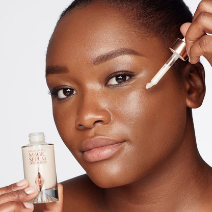 A deep-tone model with brown eyes wearing subtle eye makeup and applying a luminous face serum on her face with a gold-coloured dropper.