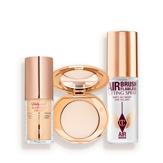 Travel-size kit that includes a glowy primer in a glass bottle with gold-coloured lid, face powder compact in a light shade with a mirrored-lid in gold-coloured packaging, and setting spray in a clear bottle with gold-coloured lid.