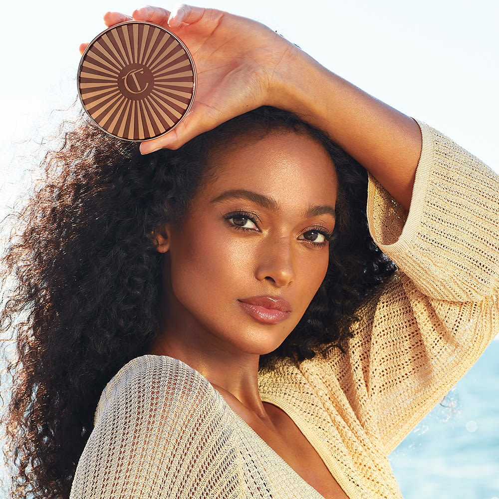 Deep-tone brunette model with glowy bronzed skin and soft-glam pink makeup holding a closed cream bronzer compact.