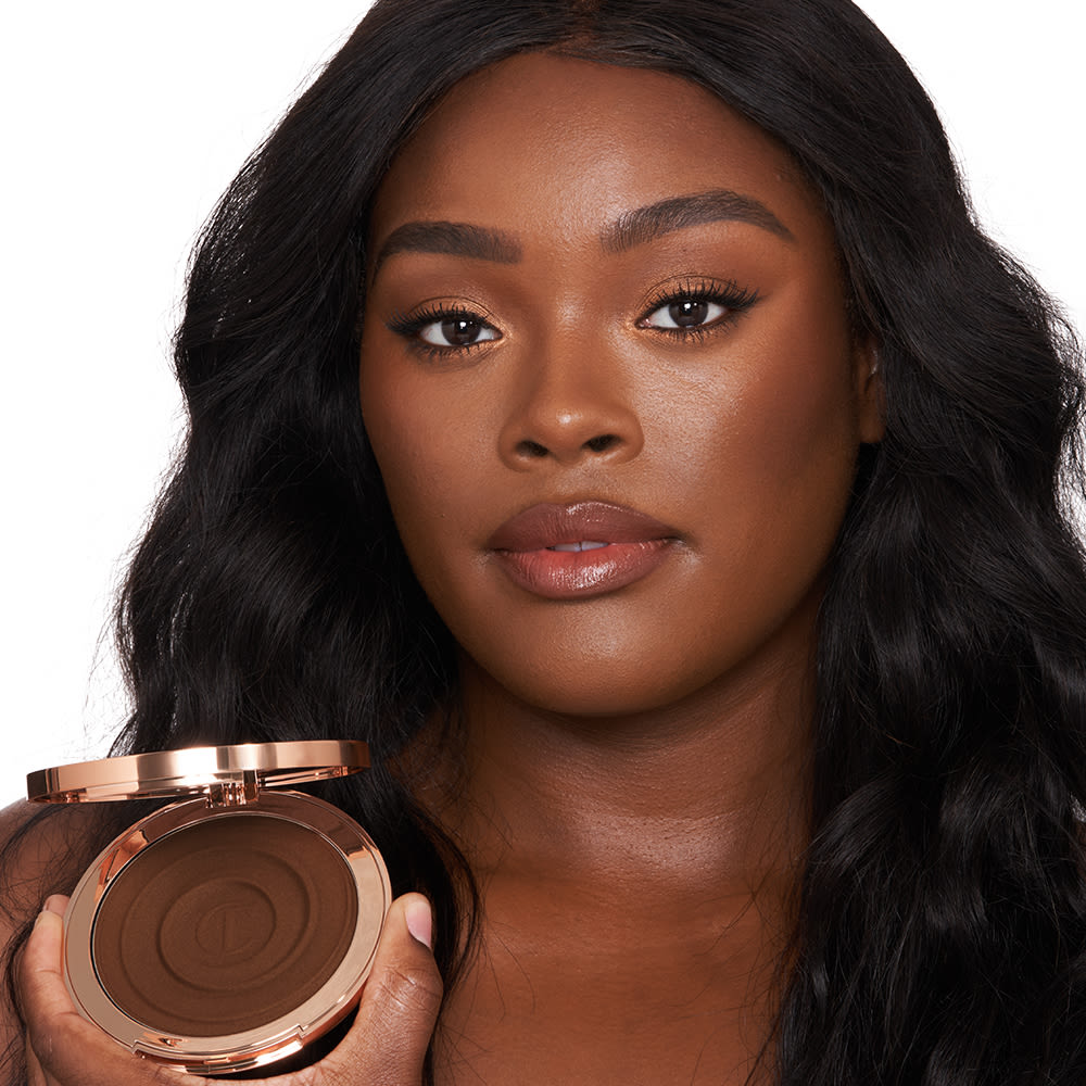 Deep-tone brunette model with brown eyes wearing nude pink lip gloss with glowy, cream bronzed for a sculpted yet natural makeup look while holding up an open bronzer compact.