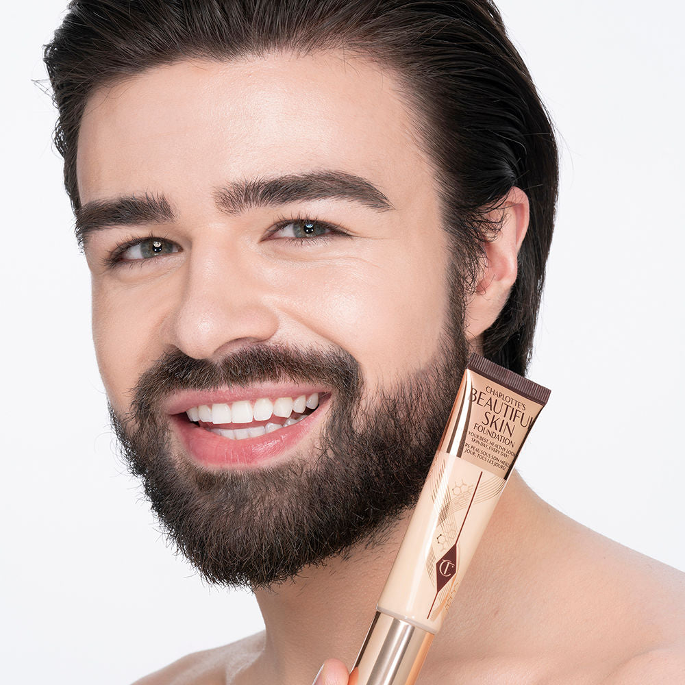 Light-tone brunette male model wearing glowy, skin-like foundation with a satin finish with nude lipstick and subtle eye makeup.