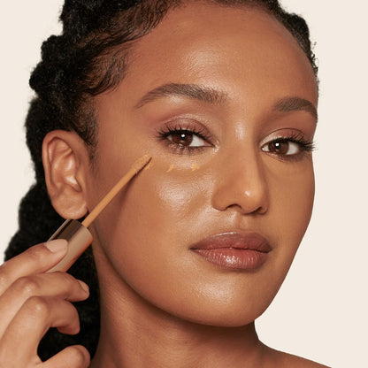 Deep-tone model wearing dewy, soft-glam makeup applying glowy, concealer in a dark brown shade.