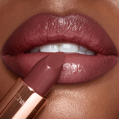 A dark, brownish-pink lipstick in front of a deep-skin model's lips that are coloured a glistening, brownish-pink.