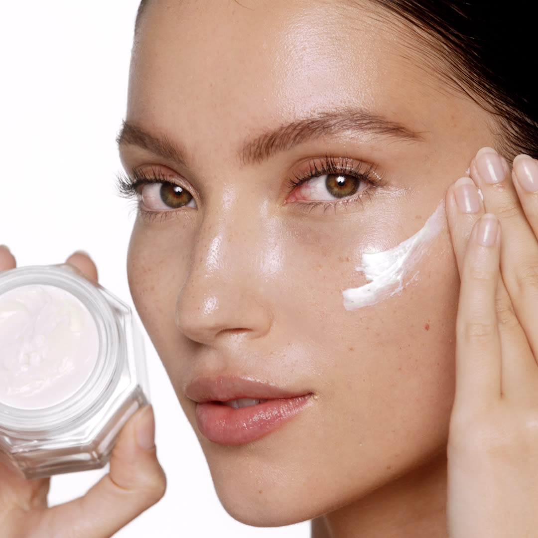 Fair-tone brunette model with glowy, flawless, glass skin, holding an open jar of pearly-white face cream and applying the cream on her face.