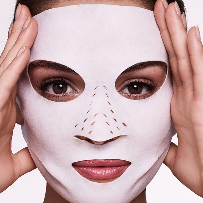 Close-up of a light-tone, brunette model wearing a sheet mask made of textile fabric with eyes, lips, and nose openings.