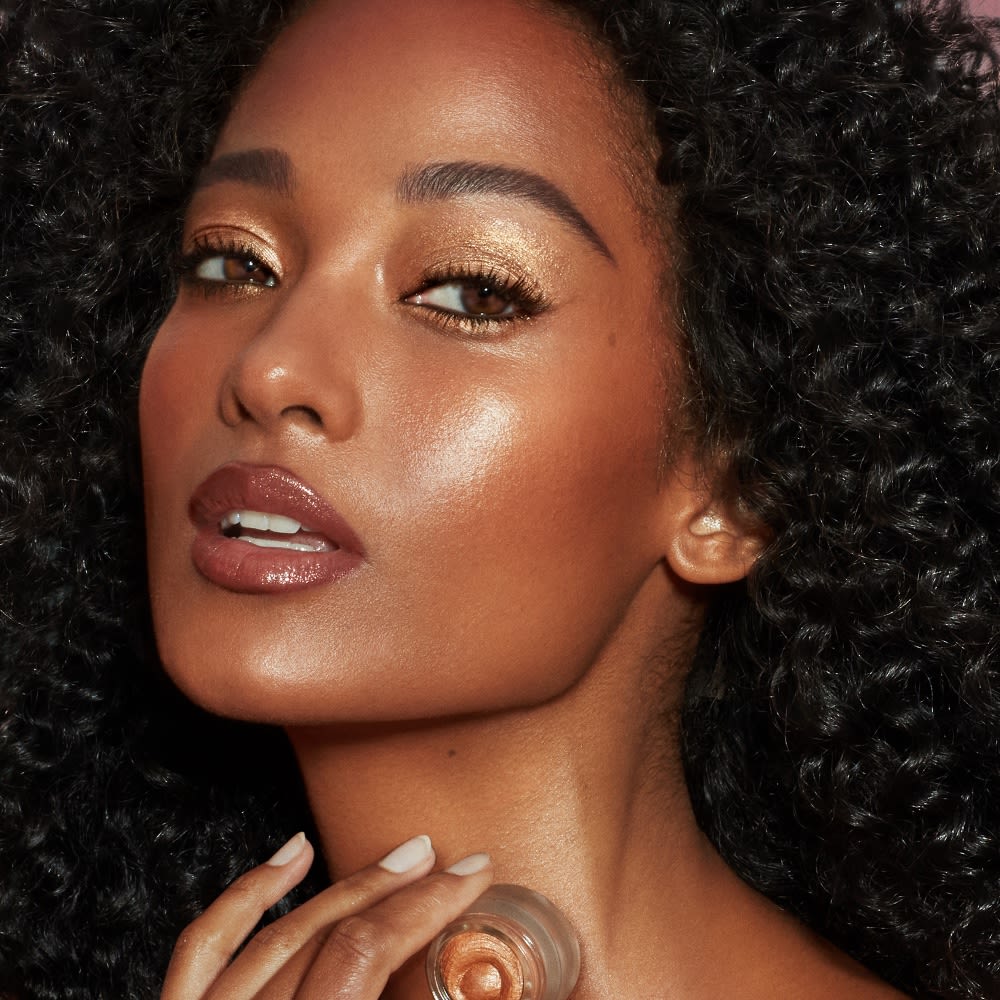 A deep-tone model with brown eyes wearing warm sunset gold cream eyeshadow with nude brown-pink lip gloss.