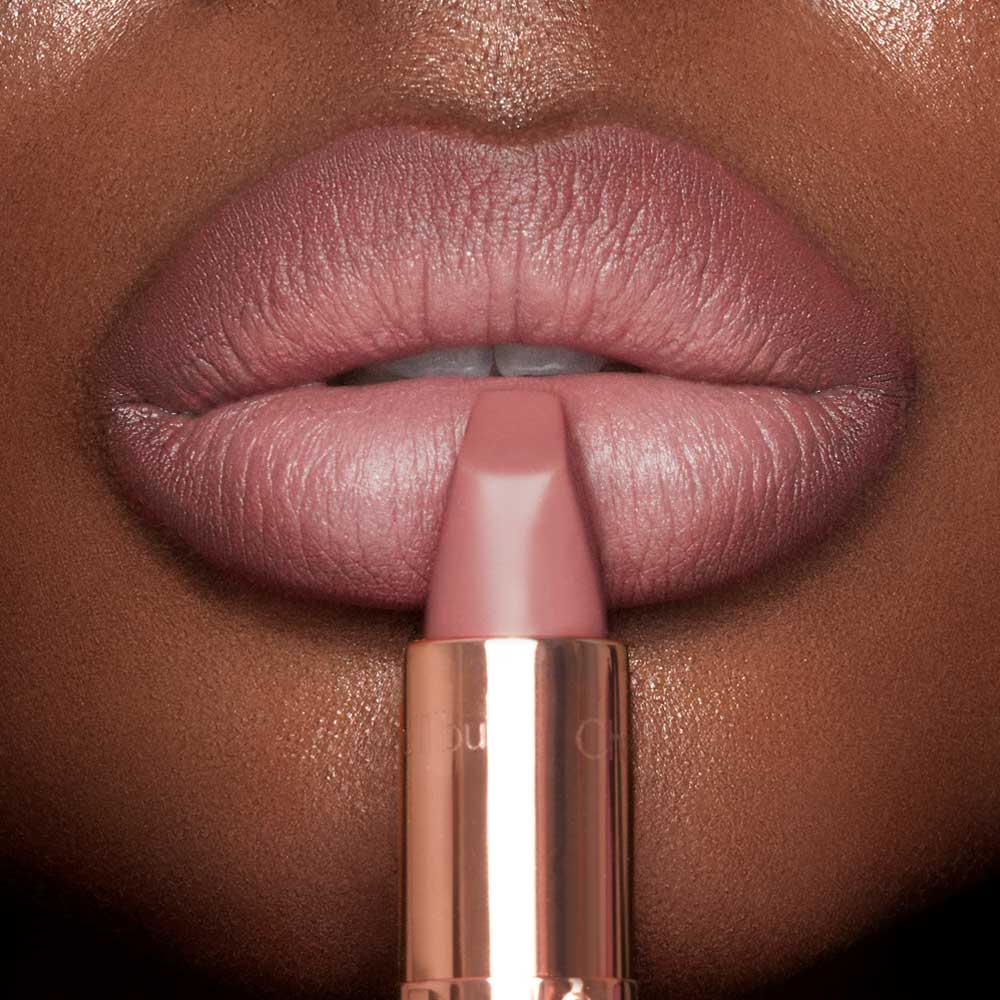 A deep-skin model wearing a nude-pink lipstick and holding the lipstick, gently pressed to their lower lip.