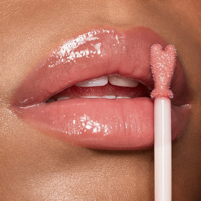 Lips close-up of a light-tone model applying sheer, nude pink lip gloss with a heart-shaped applicator.