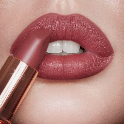 Lips close-up of a light-tone model wearing a warm, berry-pink matte lipstick while holding the lipstick in front of her lips.