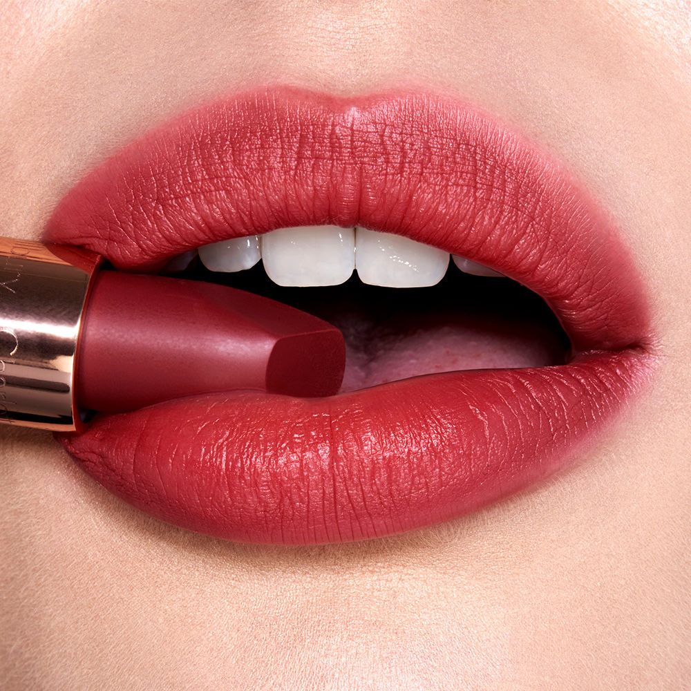 Lips close-up of a fair-tone model wearing a cranberry-red lipstick while holding the lipstick in her mouth.