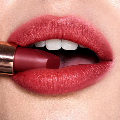 Lips close-up of a fair-tone model wearing a cranberry-red lipstick while holding the lipstick in her mouth.