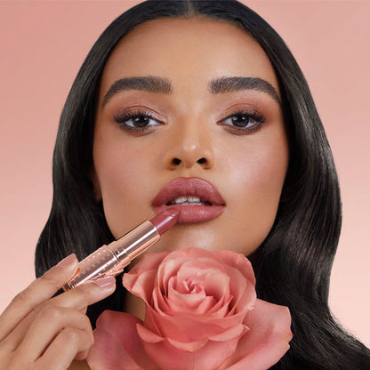 A medium-tone brunette model with brown eyes applying a matte lipstick in a golden peachy-pink shade.