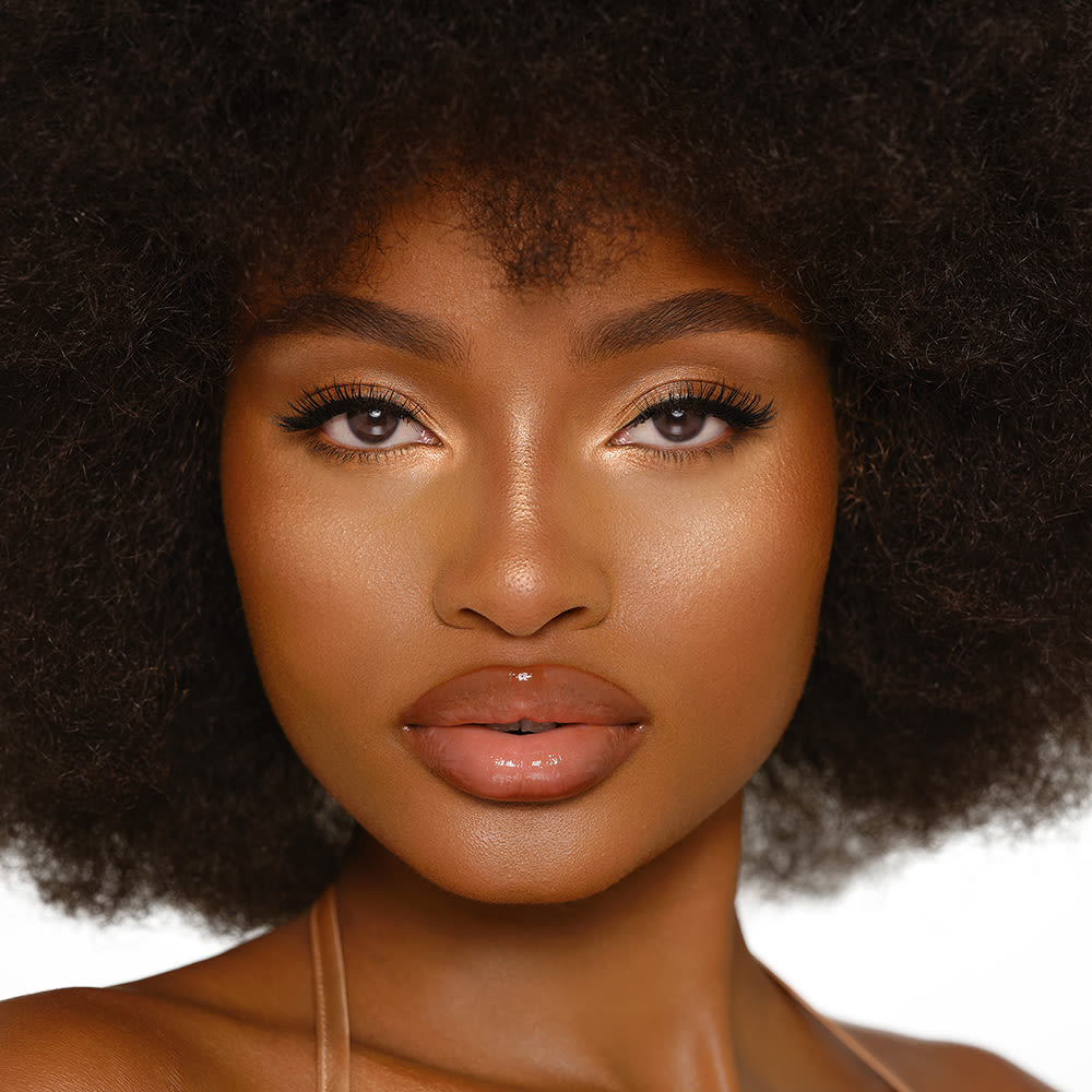 Deep-tone brunette model wearing glowy highlighter in a copper-gold shade with nude pink lip gloss and smokey eye makeup.