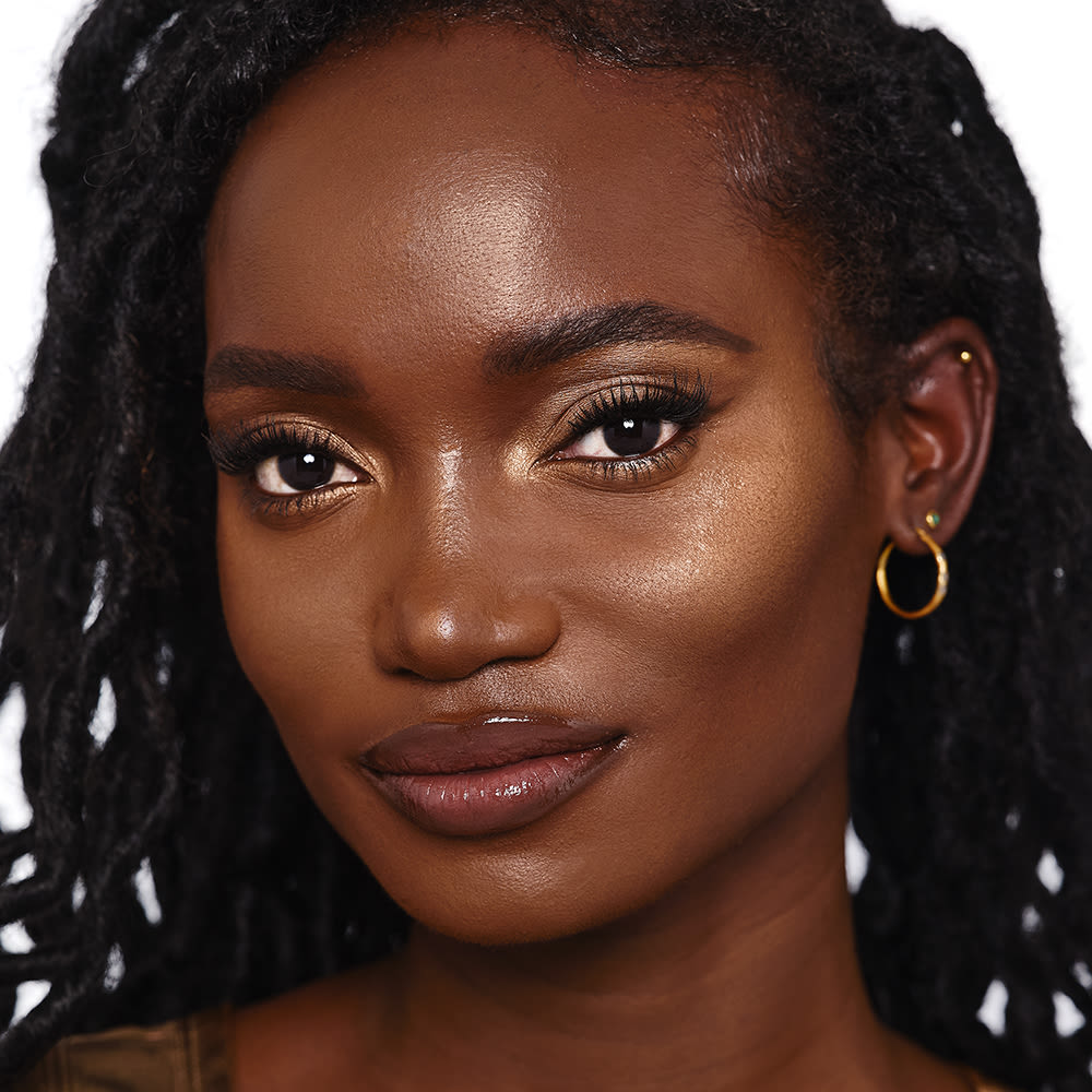 Deep-tone brunette model wearing glowy highlighter in a bronze-gold shade with vampy-purple lip gloss and smokey eye makeup.
