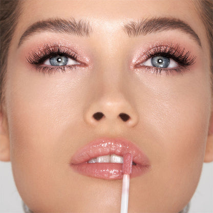 Fair-tone model with blue eyes wearing shimmery rose gold eye makeup and applying a pink glittering lip gloss with rose gold sparkle.