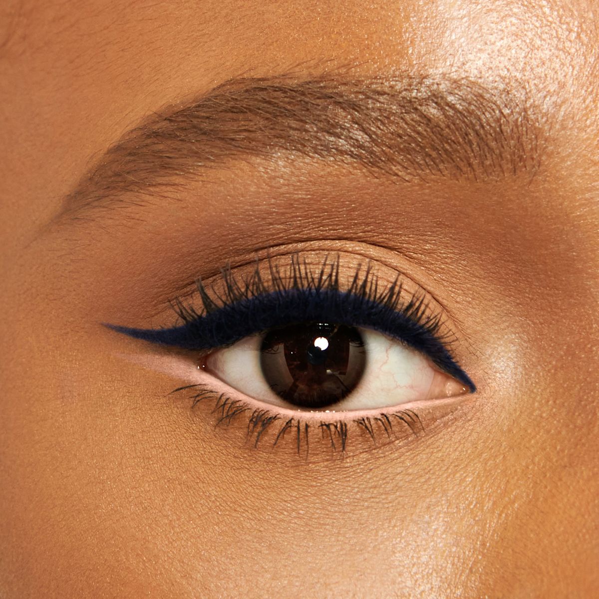 Close-up of a deep-tone model with brown eyes wearing jet-black eyeliner pencil on her eyelid and nude beige eyeliner pencil on her lower waterline.