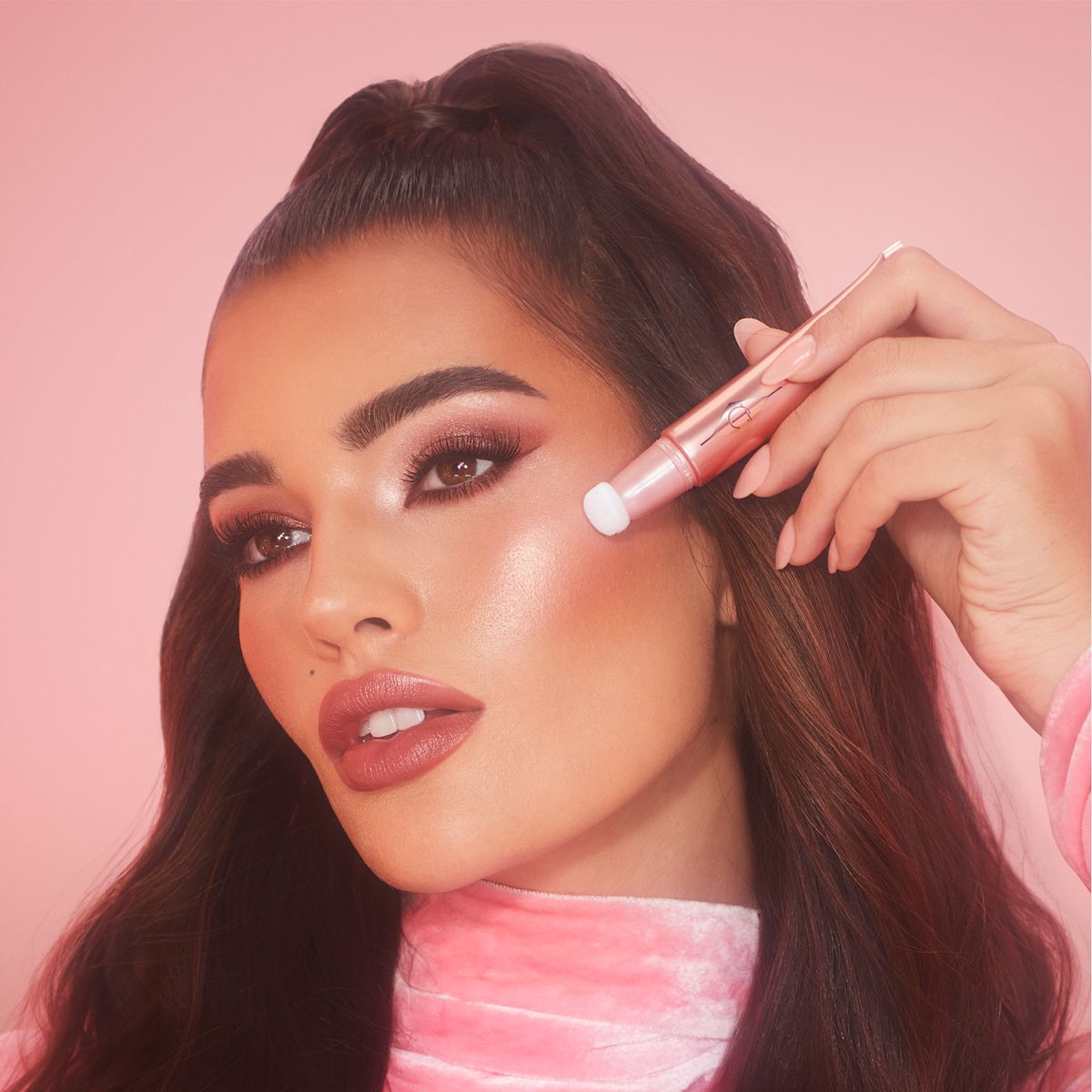 Deep-tone brunette model wearing glowy, nude pink, full-glam makeup and applying pink-peach liquid highlighter-blush from a highlighter blush wand with a soft-sponge applicator end.