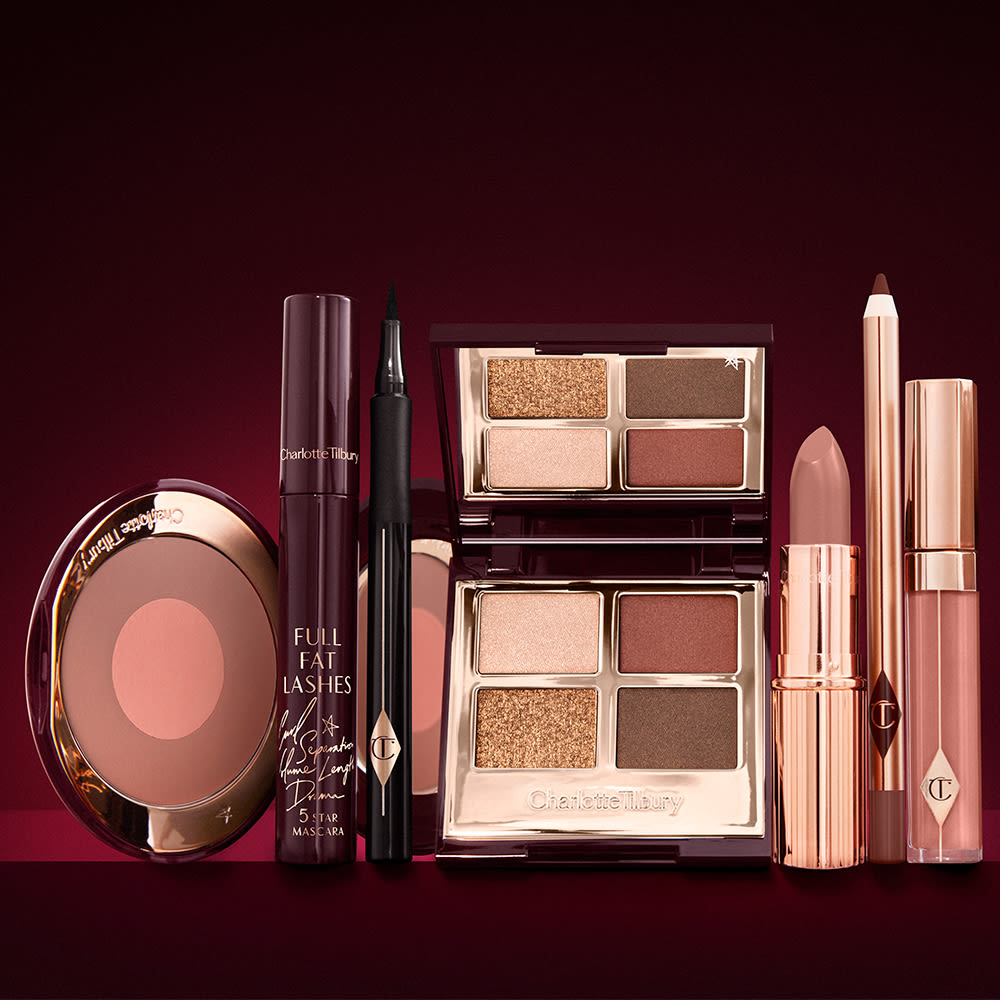 An open two-tone blush in cool-toned brown and warm pink with a mascara, eyeliner pen, quad eyeshadow palette with shimmery and matte brown and golden shades, an open lipstick in nude peach, lip liner pencil in taupe-brown, and a lip gloss in nude pink.