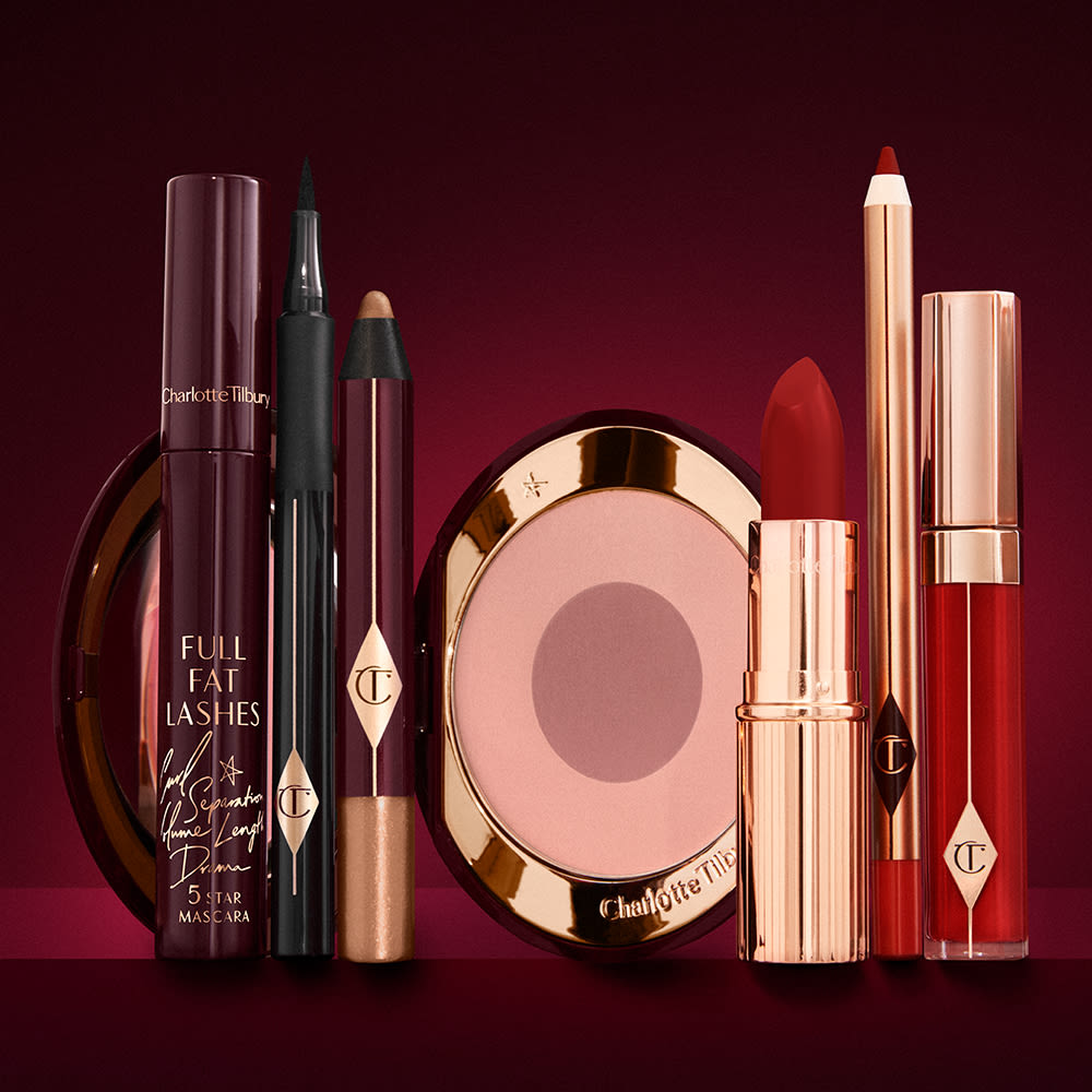 An open two-tone blush in cool-toned pink and brown with a mascara, eyeliner pen, chubby eyeshadow stick in gold, an open lipstick in bold red, lip liner pencil in blood-red, and a lip gloss in bright red.