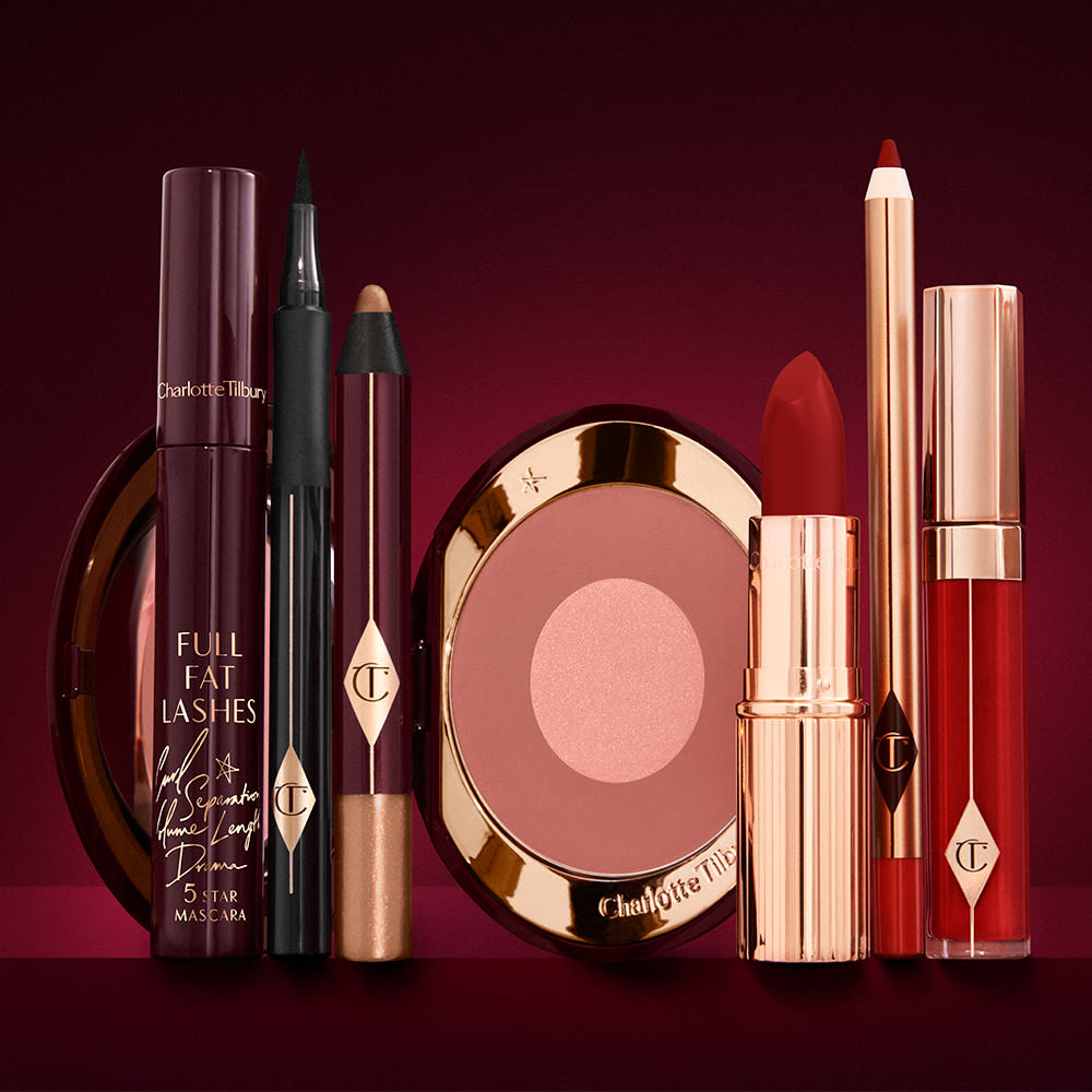 An open two-tone blush in cool-toned terracotta and warm pink with a mascara, eyeliner pen, an open lipstick in bold red, lip liner pencil in blood-red, and a lip gloss in bright red.