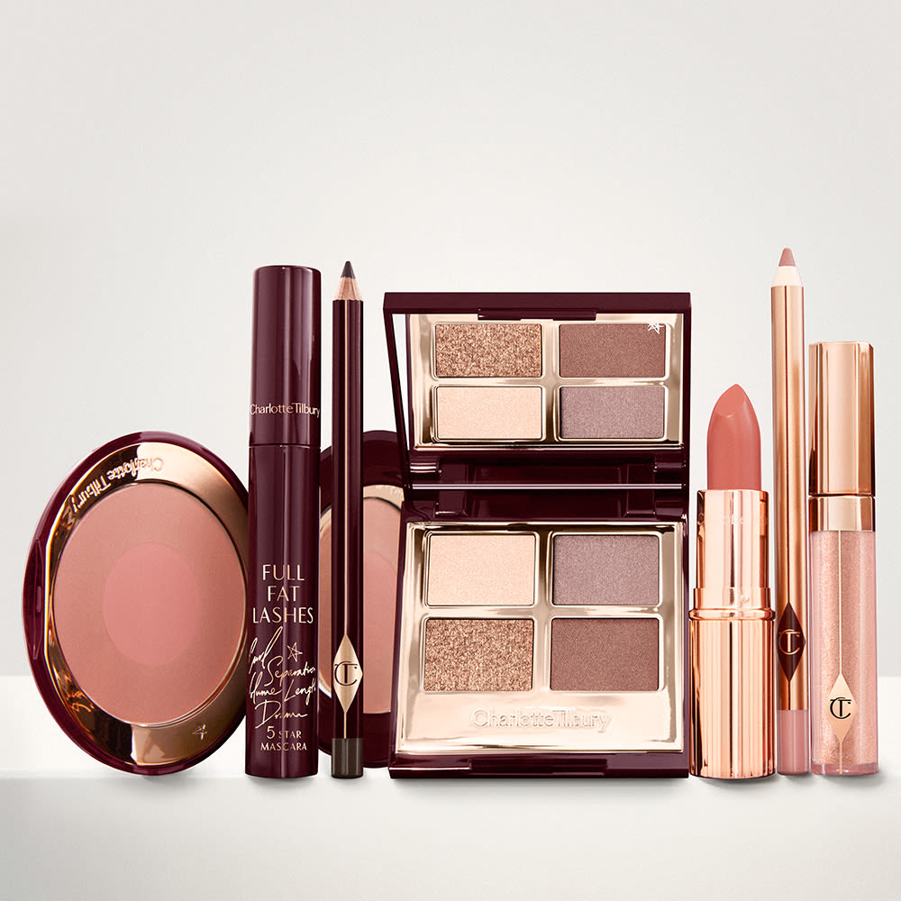 An open, mirrored-lid eyeshadow palette in matte and shimmery gold, brown, and beige shades, an open black eyeliner pencil, a mascara in a dark-crimson colour scheme, a golden-peach lipstick with a matching lip liner pencil, nude-peach lip gloss, and an open two-tone blush in muted brown-pink.