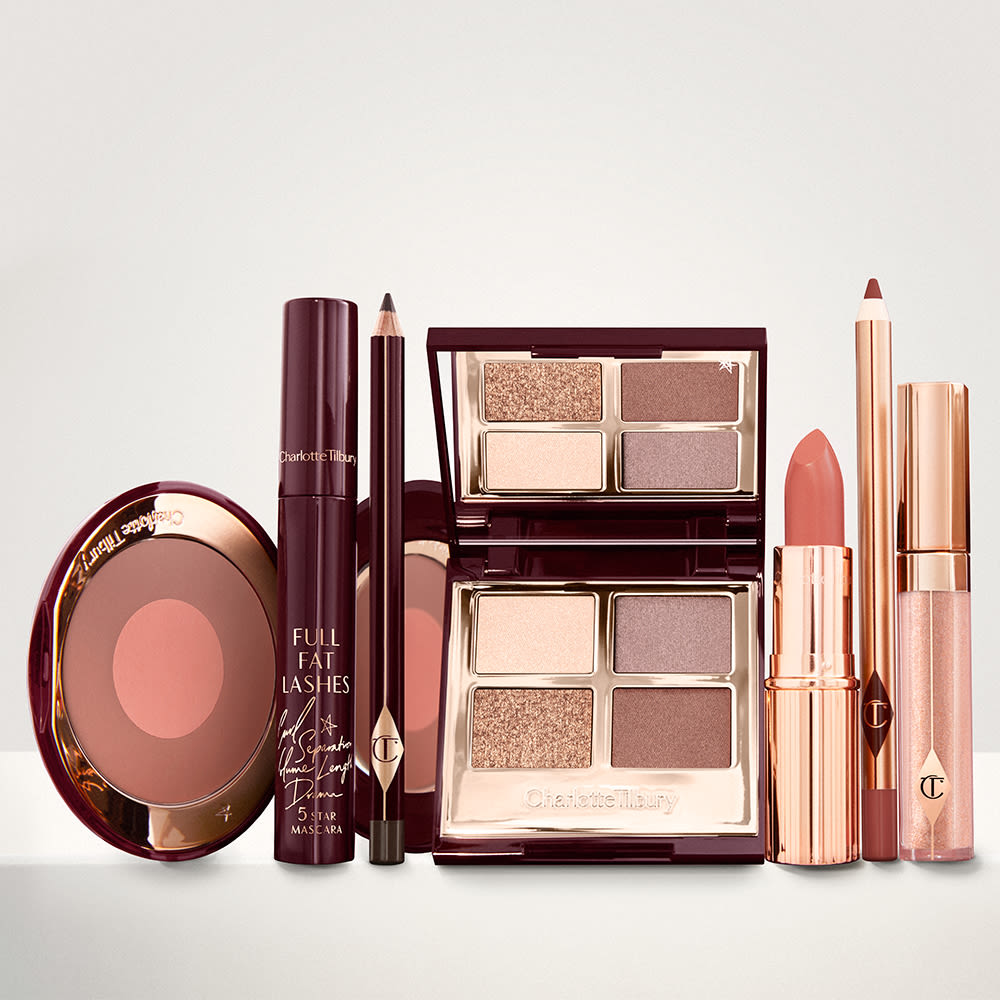 An open two-tone blush in cool-toned brown and warm pink with a mascara, eyeliner pencil, quad eyeshadow palette with shimmery and matte brown and golden shades, an open lipstick in warm pink, lip liner pencil in maroon, and a lip gloss in nude pink.