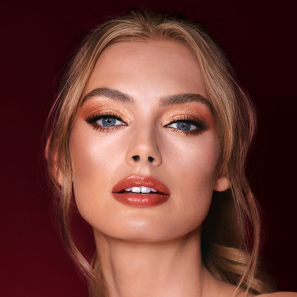 A light-tone model with blue eyes wearing shimmery copper and gold eye makeup with black eyeliner, glowy bronzed cheeks, and orange-red lipstick with gloss on top.