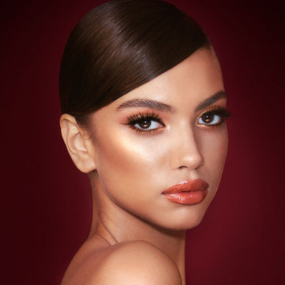 A medium-tone model with brown eyes wearing shimmery copper and gold eye makeup with black eyeliner, glowy bronzed cheeks, and orange-red lipstick with gloss on top.