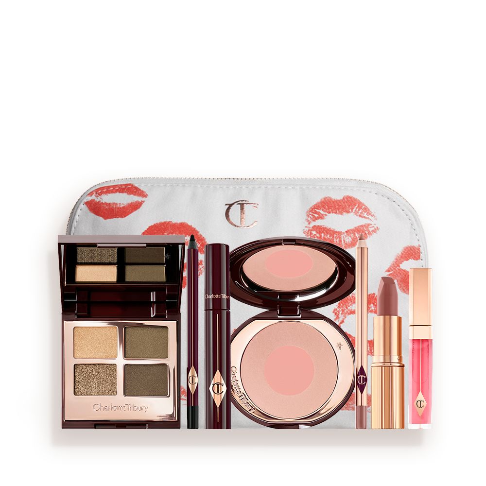 A white makeup bag with a quad eyeshadow palette in shades of green and gold, a brown eyeliner pencil, mascara, an open two-tone blush in nude pink, a warm pink lip liner pencil, a warm rose lipstick, and a bright pink lip gloss.