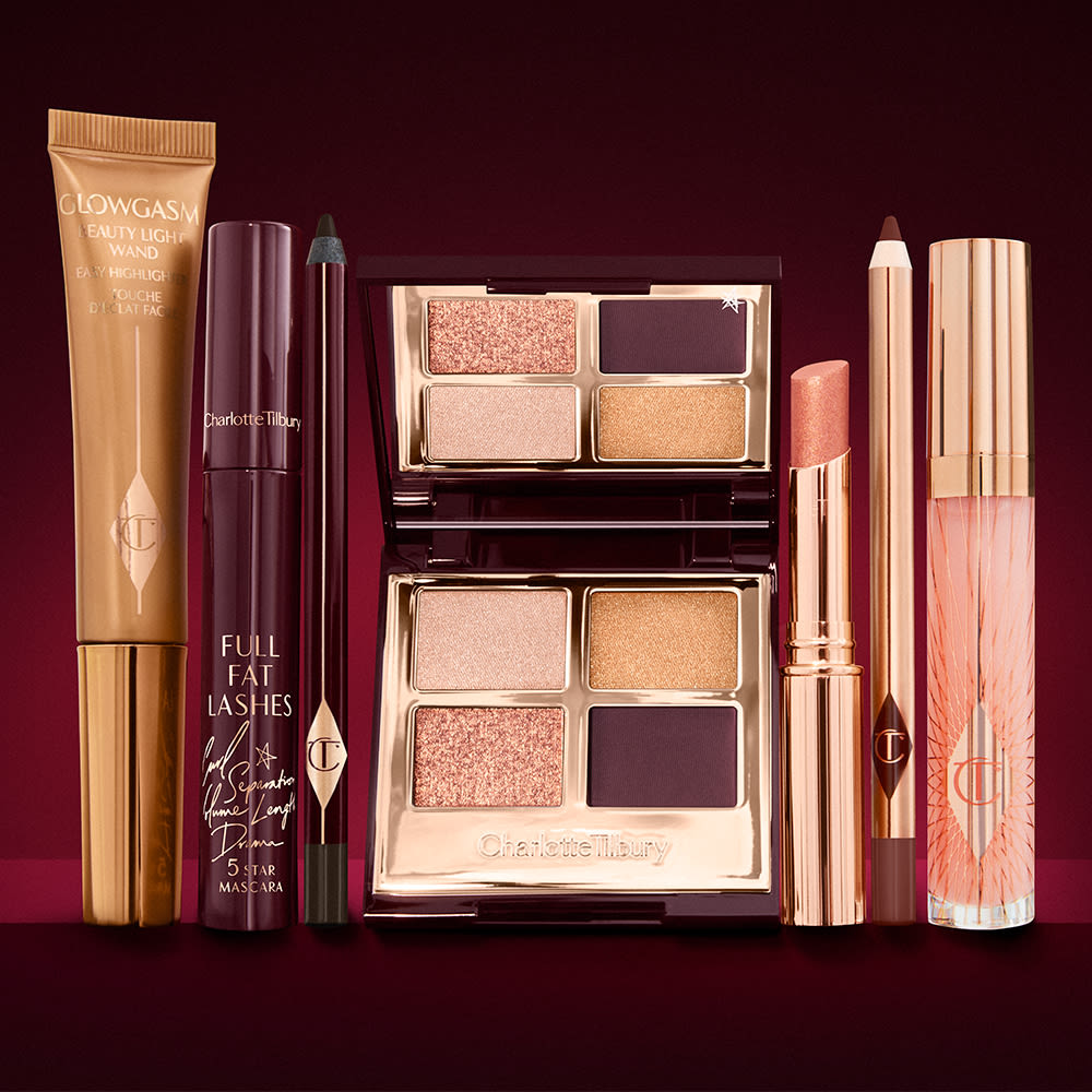 A highlighter wand in a honey-gold-coloured tube with a mascara, eyeliner pencil, quad eyeshadow palette with shimmery and matte rose gold, plum, and golden shades, an open lipstick in nude coral, lip liner pencil in vampy red, and a lip gloss in nude pink.