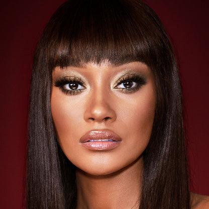 A deep-tone model with brown eyes wearing smokey green eye makeup with black eyeliner, soft brown blush, and nude pink lipstick with gloss on top.