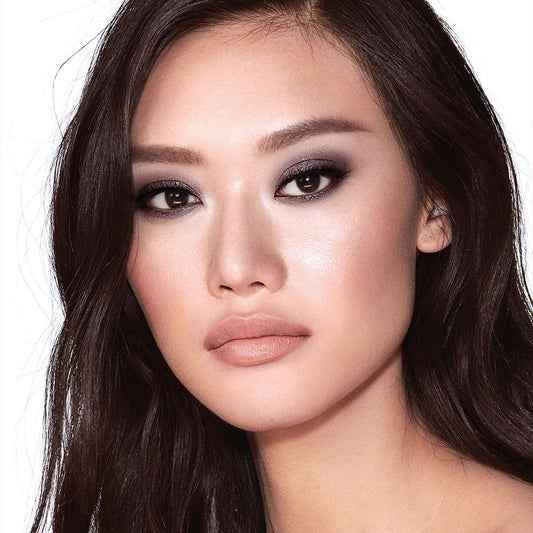 A fair-tone brunette model with brown eyes wearing smokey silvery eyeshadow with bronzed cheeks and glossy nude-peach lips.