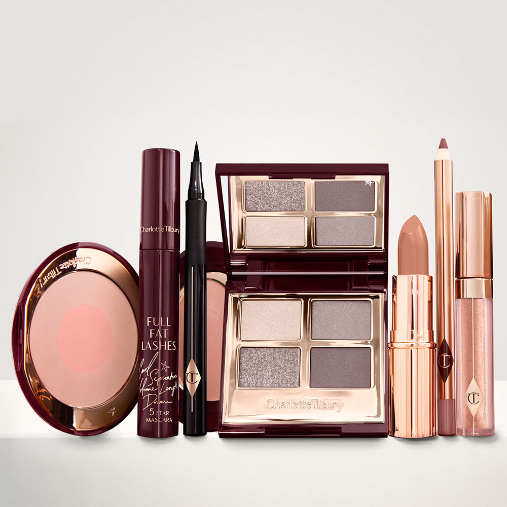 An open, mirrored-lid eyeshadow palette in matte and shimmery silver, grey, and beige shades, an open black eyeliner pen, a mascara in a dark-crimson colour scheme, a nude-pink lipstick with a matching lip liner pencil, nude pink lip gloss, and an open two-tone blush in muted rose-gold and pink.