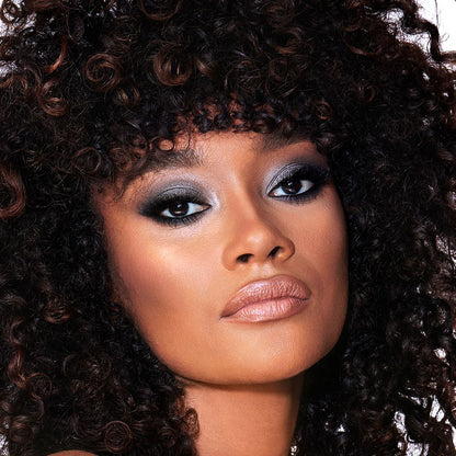 A deep-tone brunette model with brown eyes wearing smokey silvery eyeshadow with bronzed cheeks and glossy nude-peach lips.