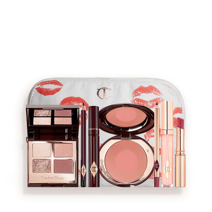 A white makeup pouch with an open two-tone blush in cool-toned brown and warm pink with a mascara, eyeliner pencil, quad eyeshadow palette with shimmery and matte brown and golden shades, an open lipstick in nude red, lip liner pencil in maroon and a lip gloss in nude pink.