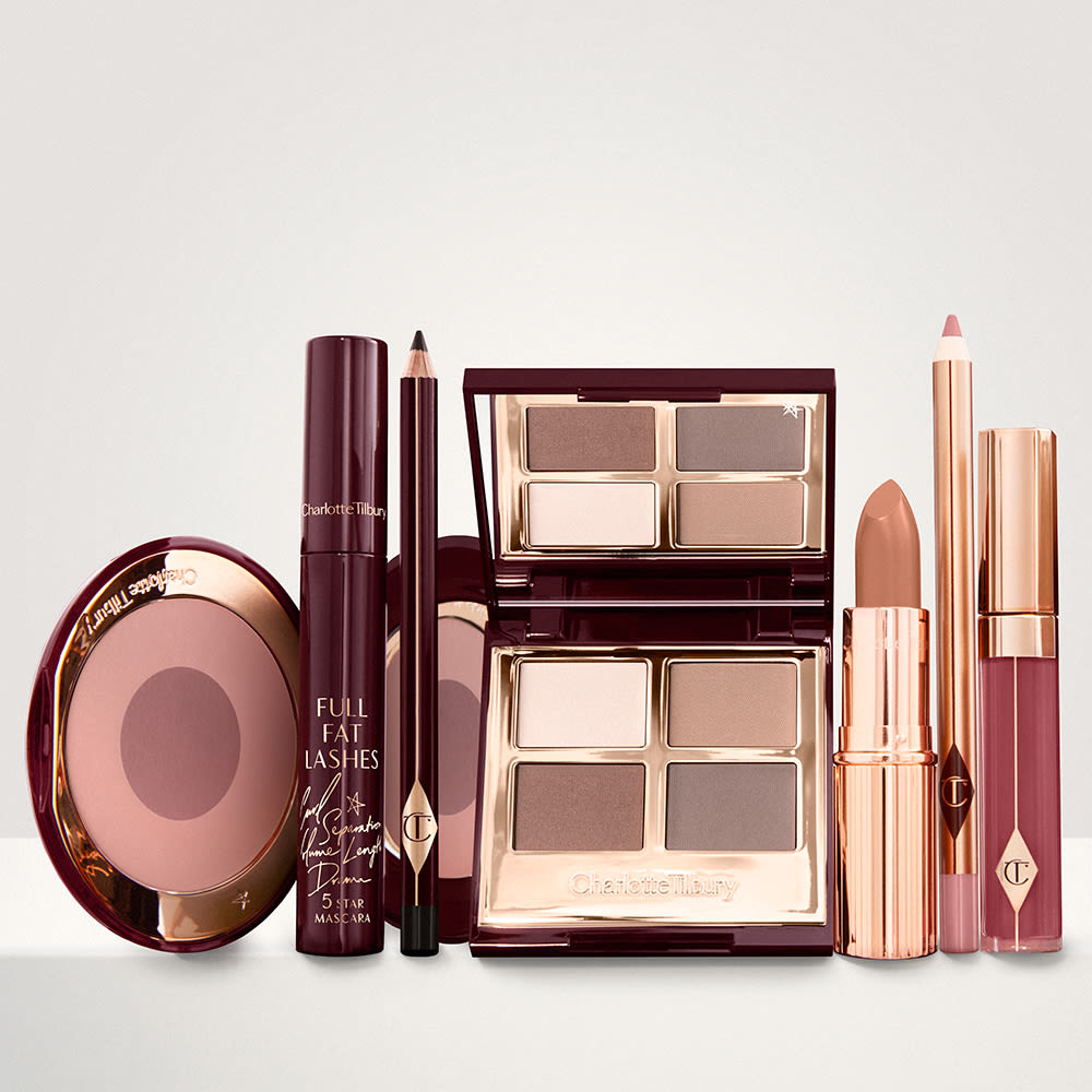 An open, mirrored-lid eyeshadow palette in matte and shimmery taupe, grey, and beige shades, an open black eyeliner pencil, a mascara in a dark-crimson colour scheme, a nude-peach lipstick with a matching lip liner pencil, magenta lip gloss, and an open two-tone blush in muted brown-pink.