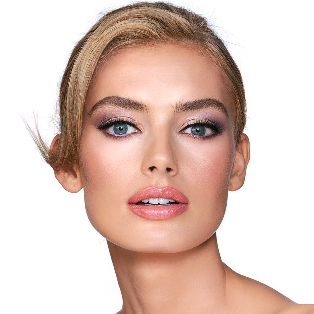 A light-tone blonde model with blue eyes wearing shimmery lilac and champagne eye makeup with nude pink blush and a warm, coral lipstick.