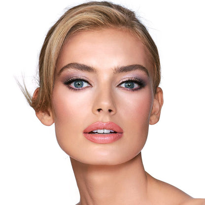 A light-tone blonde model with blue eyes wearing shimmery lilac and champagne eye makeup with nude pink blush and a warm, coral lipstick.