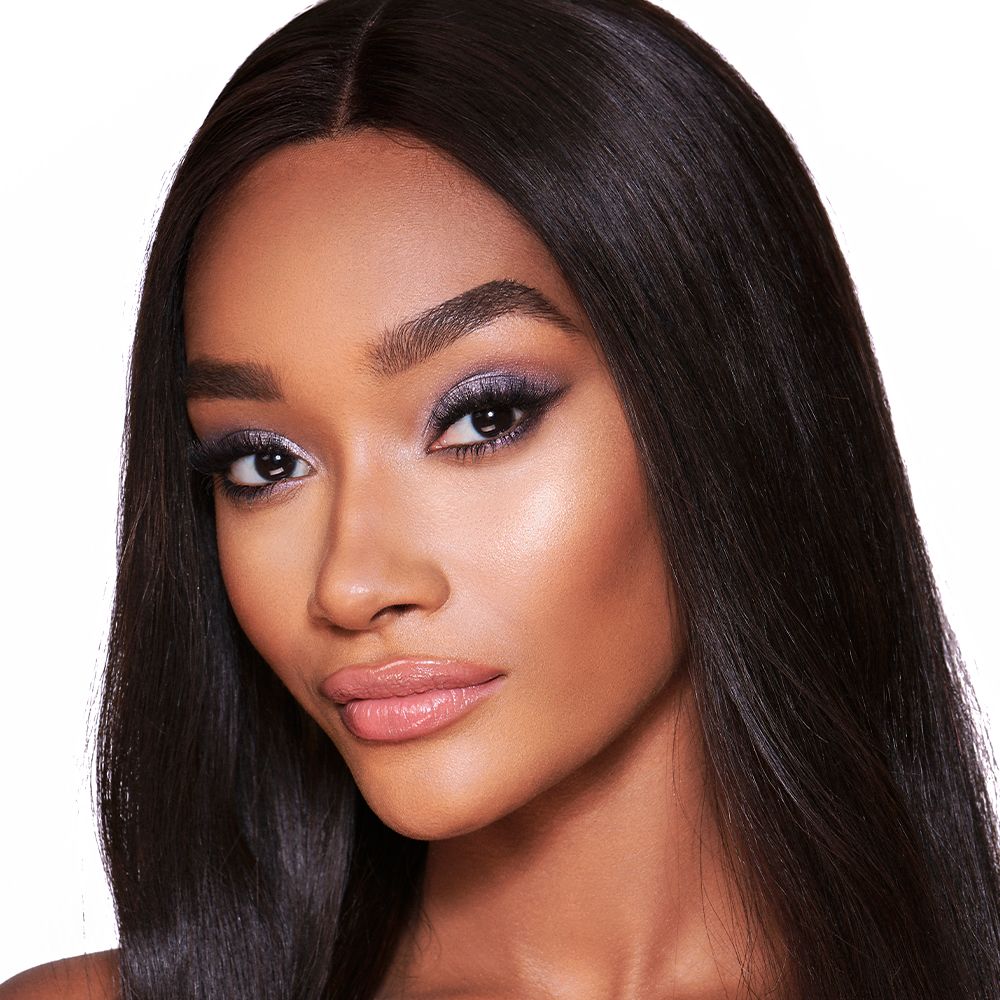 A deep-tone blonde model with brown eyes wearing shimmery lilac and champagne eye makeup with nude pink blush and a warm, coral lipstick.