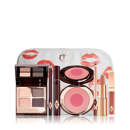 A makeup bag with 7 makeup products, an open two-tone blush in cool-toned pink and fuchsia with a mascara, eyeliner pencil, quad eyeshadow palette with shimmery and matte grey and golden shades, an open lipstick in nude red, lip liner pencil in taupe-brown, and a lip gloss in warm brown-peach.