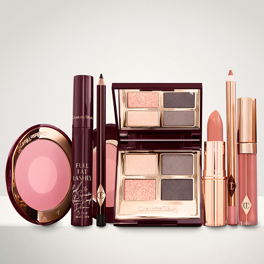 An open, mirrored-lid eyeshadow palette in matte and shimmery gold and grey shades, an open black eyeliner pencil, a mascara in a dark-crimson colour scheme, a golden-peach lipstick with a matching lip liner pencil, coral lip gloss, and an open two-tone blush in cool pink.