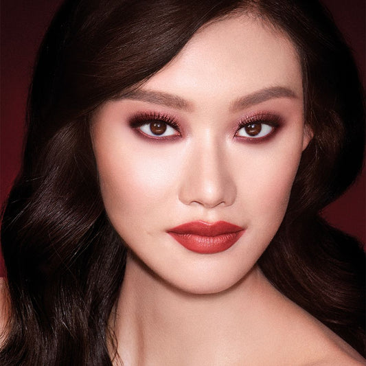 A fair-tone model with brown eyes wearing reddish-plum eye makeup, glowy face base, and a vampy-red lipstick with a satin-finish.