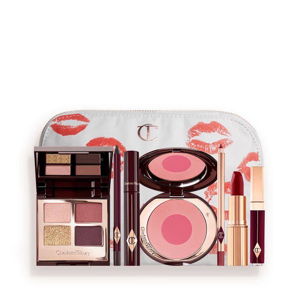A makeup bag with 7 makeup products, an open, mirrored-lid eyeshadow palette in matte and shimmery gold and red shades, an open black eyeliner pencil, a mascara in a dark-crimson colour scheme, a red lipstick with a matching lip liner pencil, vampy-red lip gloss, and an open two-tone blush in cool pink.