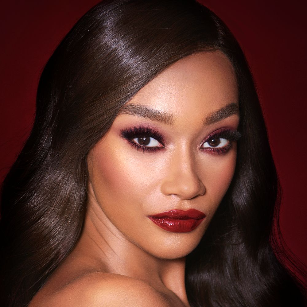 A deep-tone brunette model with reddish-plum eye makeup, glowy face base, and a vampy-red lipstick with a satin-finish.