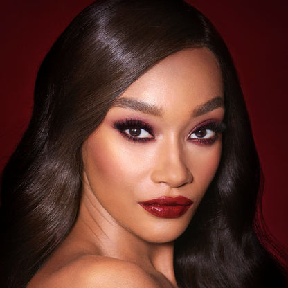 A deep-tone brunette model with reddish-plum eye makeup, glowy face base, and a vampy-red lipstick with a satin-finish.