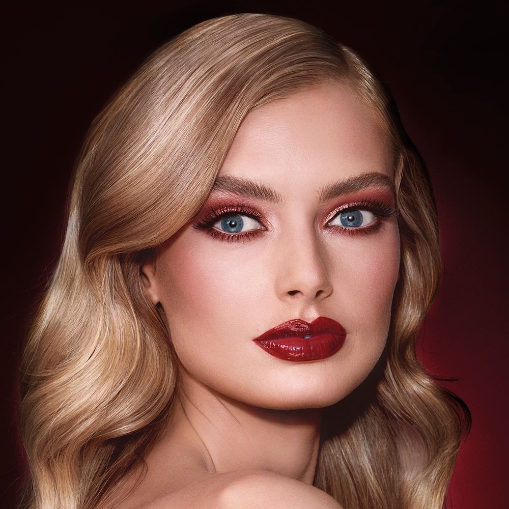 A fair-tone blonde model with reddish-plum eye makeup, glowy face base, and a vampy-red lipstick with a satin-finish.