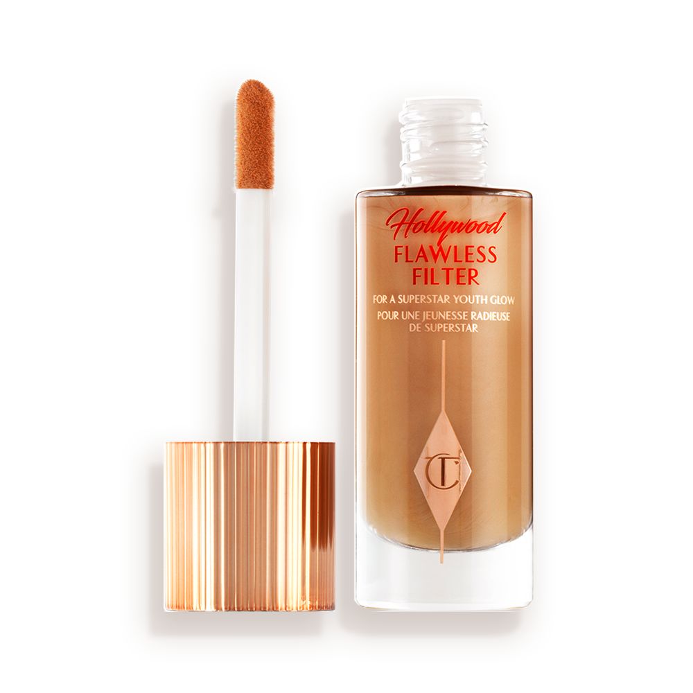 An open, luminous primer in a dark golden-beige shade in a glass bottle with its gold and white doe-foot applicator next to it.